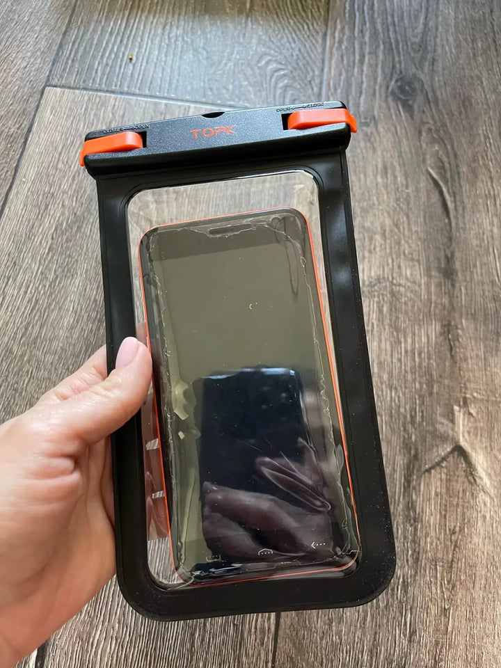 A hand holding an IPX8 Waterproof Phone Pouch, which encloses a black smartphone with an orange and black clasp, against a wooden floor background. Perfect for outdoor activities, this case offers top-notch protection.