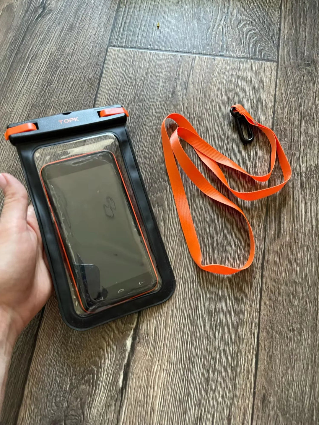 A hand holds an IPX8 Waterproof Phone Pouch equipped with an orange lanyard and carabiner on the wooden floor, ready for any outdoor activities.