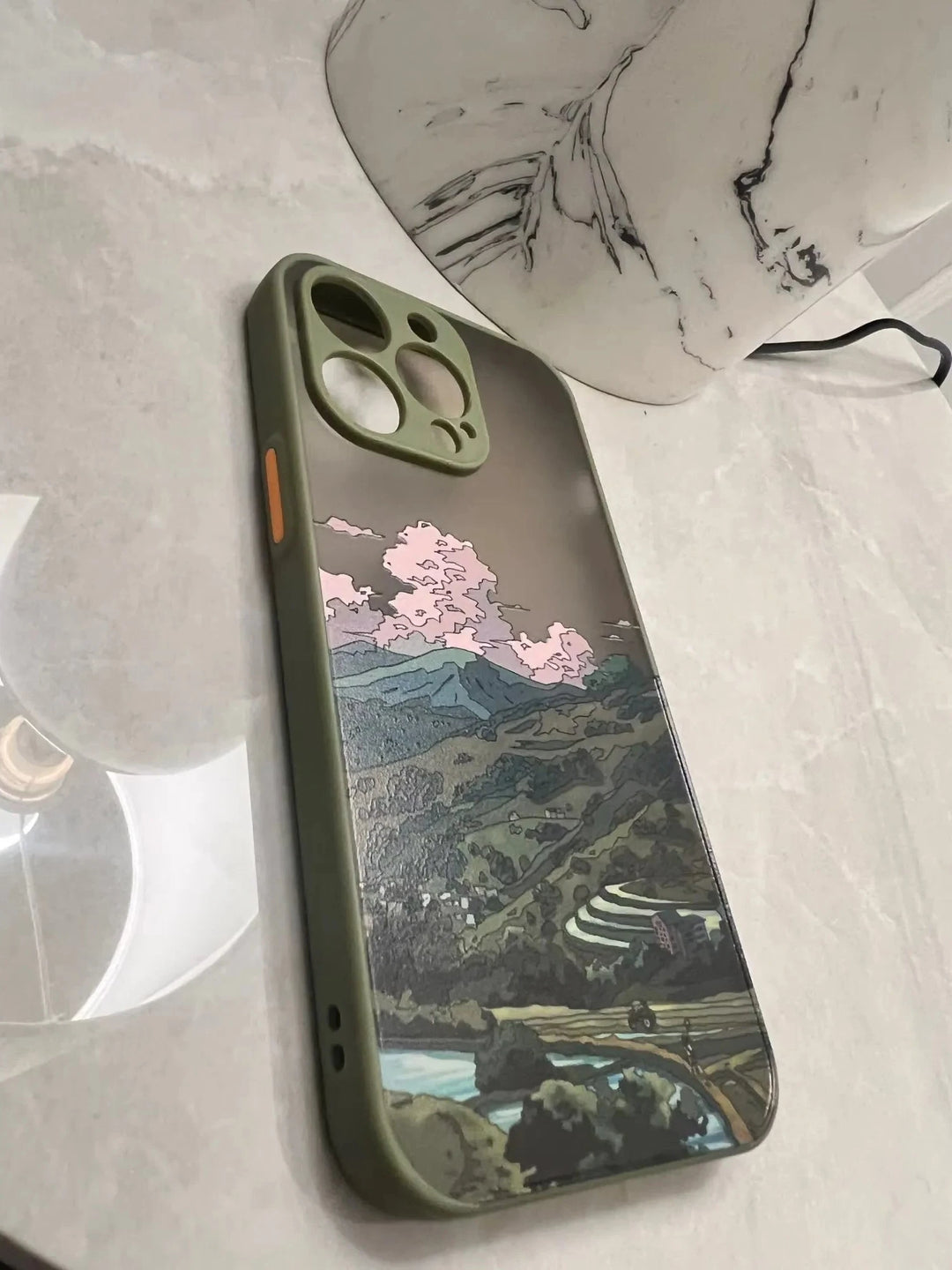 A green phone case with a frosted back featuring a landscape painting of rolling hills, a village, and a road