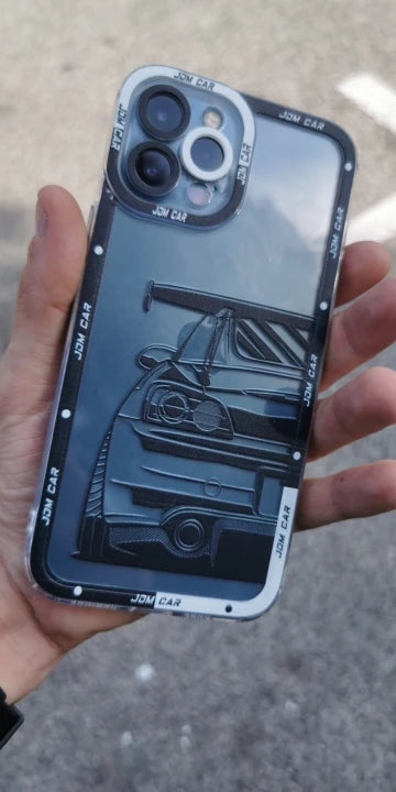 Person holding the JDM Clear for iPhone encased in a silicone case featuring a printed design of a car's rear view with a spoiler. The anti-scratch border has "JDM CAR" text. The background reveals an outdoor, concrete surface.