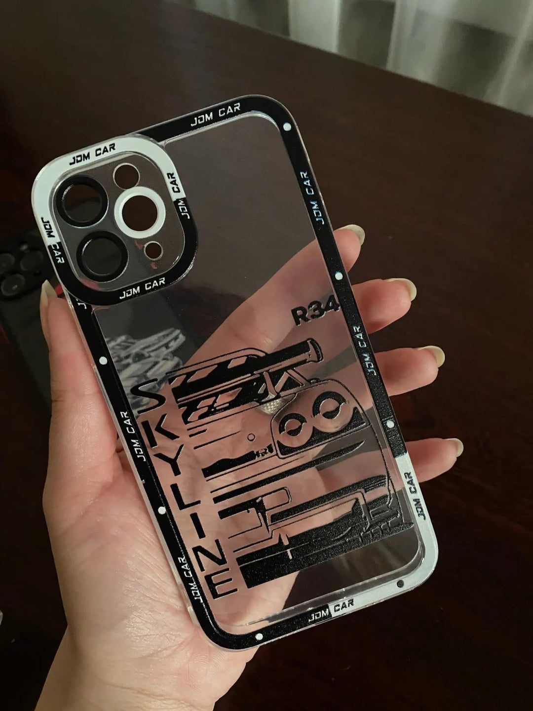 A hand holds a JDM Clear for iPhone case, which is an anti-scratch, transparent silicone case with black trim. It features JDM car designs showcasing parts of a car and the word "SKYLINE" along with the text "R34".