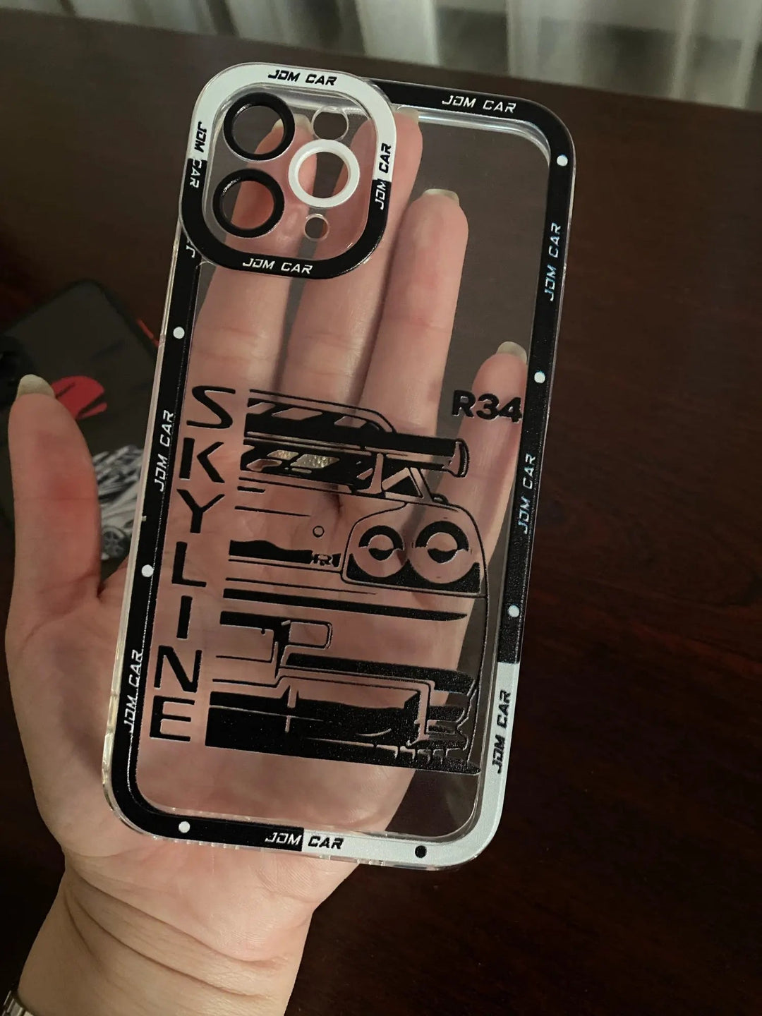 Close-up of a hand holding the JDM Clear for iPhone, a transparent silicone case adorned with the text "SKYLINE" and "R34," as well as images of car parts. This anti-scratch phone case showcases iconic JDM car designs.