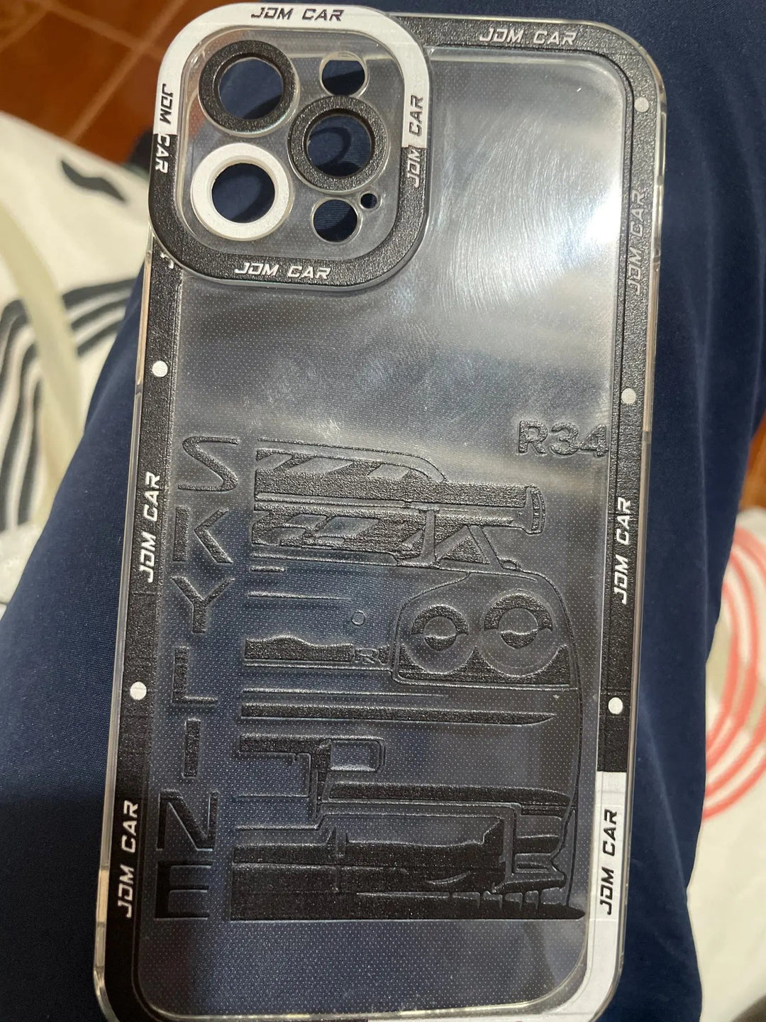 The JDM Clear for iPhone is a clear silicone case adorned with the engraved text "SKYLINE GTR R34" and a vector outline of a car. It features anti-scratch protection and has "JDM CAR" repeatedly written along the border.