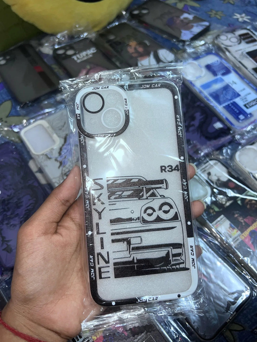 A hand holding a "JDM Clear for iPhone" silicone case featuring "Skyline R34" and JDM car designs, displayed among various other anti-scratch phone cases on a table.