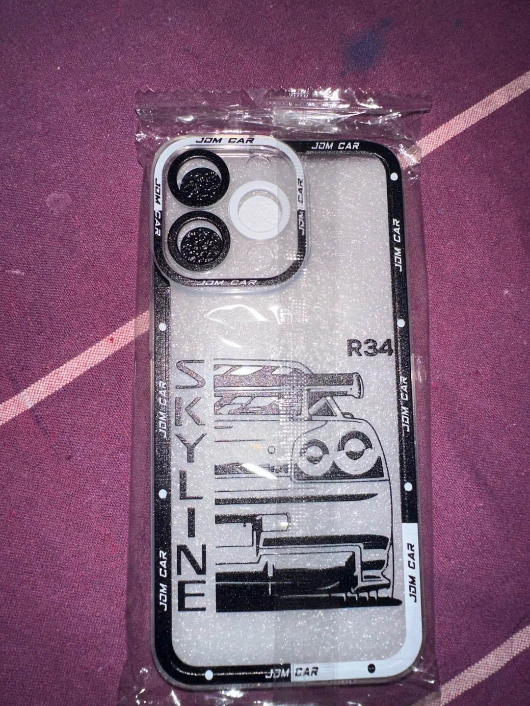 The JDM Clear for iPhone is a sleek smartphone case featuring a black and white design of a JDM car with the word "SKYLINE R34" on it, crafted from anti-scratch silicone and packaged in clear plastic.