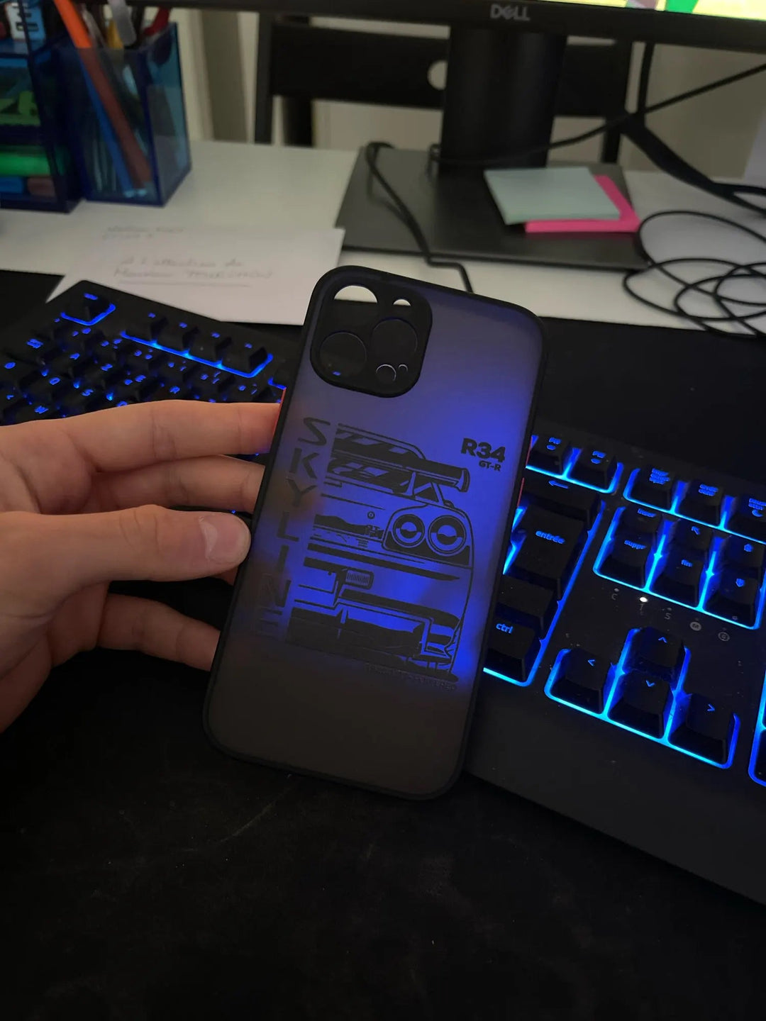 A person's hand holding the JDM Frosted for iPhone case, featuring a graphic design of a car with the text "SKYLINE" and "R34 GT-R," in front of a keyboard with blue backlighting and a computer monitor.