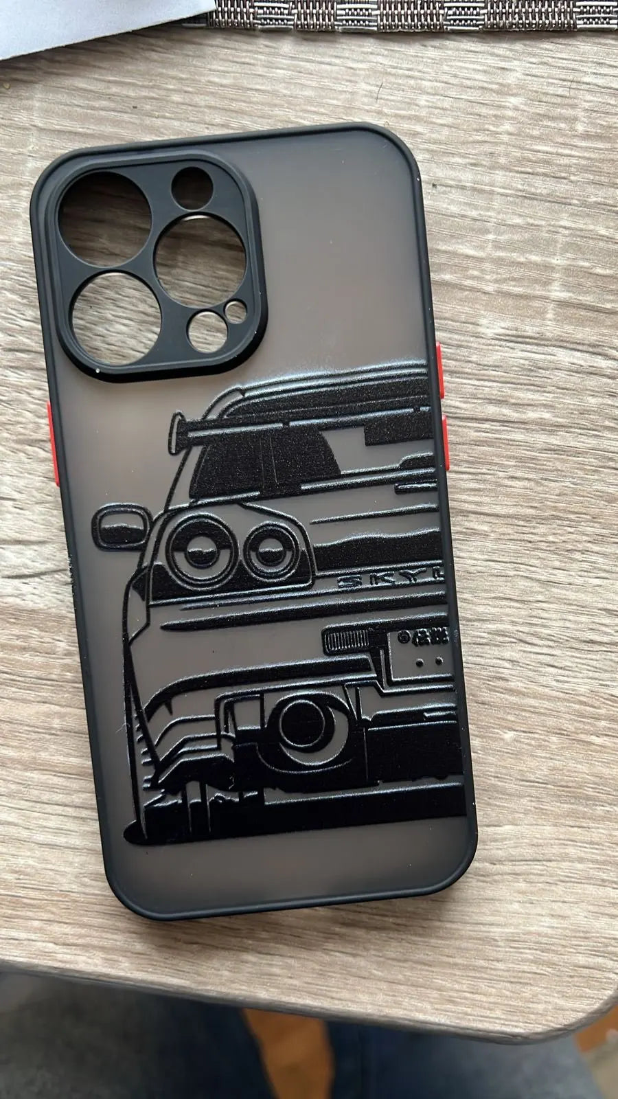 A JDM Frosted for iPhone case, featuring a black and white illustration of the rear view of a sports car, is placed on a wooden surface. This phone case not only showcases sleek automotive art but also offers robust protection for your device.