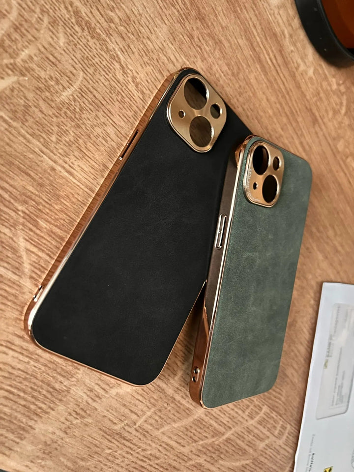 Two Lambskin Leather for iPhone cases with metallic edges and premium lambskin leather back are placed on a wooden surface. One case is black and the other is blue-gray, both featuring a gold camera cutout.