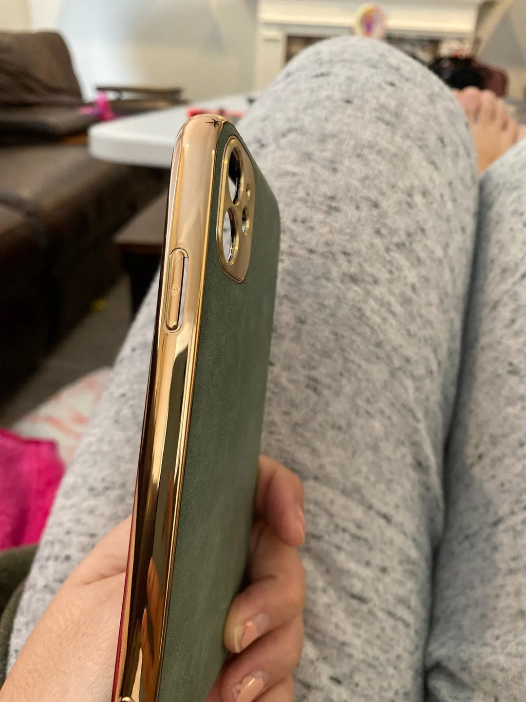 Close-up of a hand holding a smartphone protected by the Lambskin Leather for iPhone case, showcasing a gold camera cutout, resting on someone's legs covered in grey sweatpants. In the background, there's a couch and various household items.