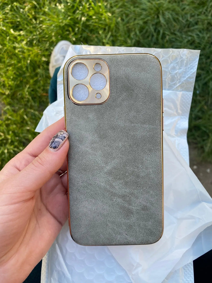 A hand holds the Lambskin Leather for iPhone, featuring luxurious gray lambskin leather and a gold camera cutout, with bubble wrap packaging underneath. Grass is visible in the background.