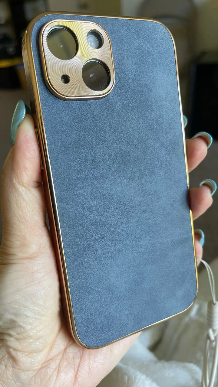 A hand holding a smartphone with the Lambskin Leather for iPhone, featuring a premium phone case in blue and gold, showcasing the phone's dual camera and flash through a sleek gold camera cutout.