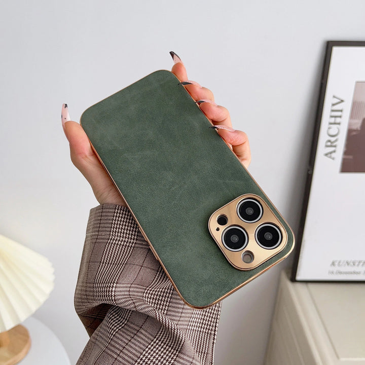 A hand holding an iPhone encased in Lambskin Leather with a gold camera cutout. The person is wearing a plaid sleeve. A framed picture and a lamp are visible in the background.