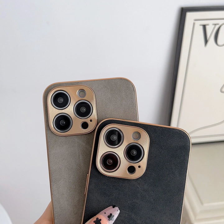 Two iPhones with camera lenses visible on the back, each elegantly encased in a Lambskin Leather for iPhone case. One phone is held with a hand displaying floral nail art, and a piece of art with text is partially visible in the background. The gold camera cutout adds a touch of sophistication to the scene.