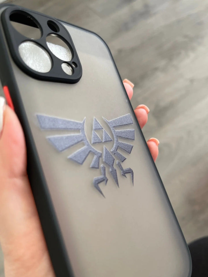 A close-up of a hand holding the "Legend of Zelda for iPhone" smartphone, adorned with a black case featuring a silver Wingcrest emblem from The Legend of Zelda series. The phone, evoking the sleek design of the Master Sword, is equipped with three camera lenses.