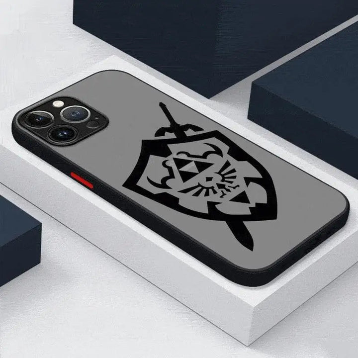 The Legend of Zelda for iPhone, which features a black case adorned with a stylized Hylian shield and sword design with a central triangular emblem reminiscent of The Legend of Zelda series, is placed on a gray surface surrounded by dark blocks.