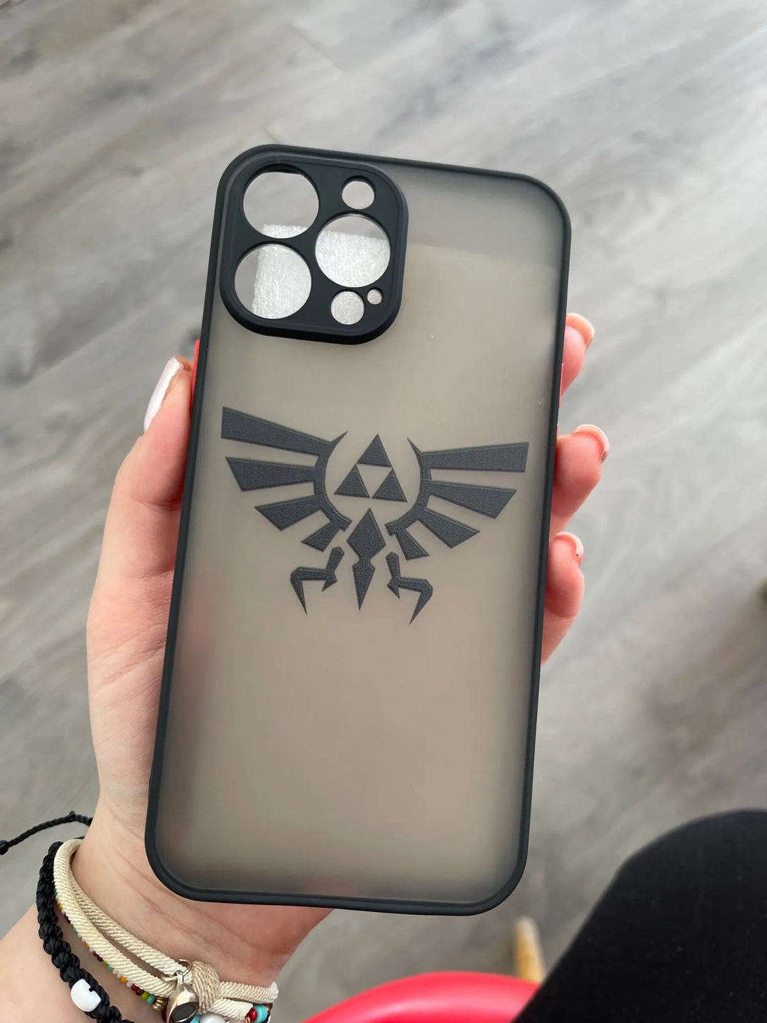 Hand holding a smartphone with the Legend of Zelda for iPhone case, featuring a black Triforce emblem reminiscent of the Hylian Shield, in front of a wooden floor background.