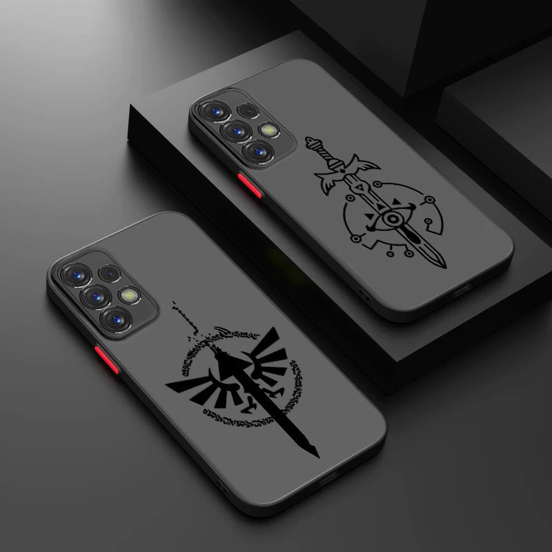 Two Legend of Zelda smartphone cases for Samsung lying on a dark surface. Each case has a distinct design: the top case features a sword reminiscent of the Master Sword with an eye symbol, while the bottom case showcases a winged bird emblem surrounded by decorative elements. Both cases are grey with black designs.