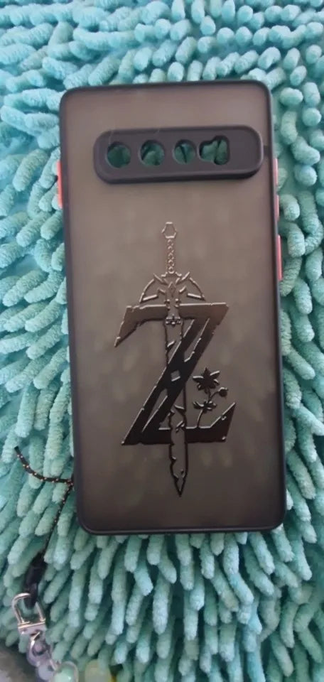 A Legend of Zelda for Samsung black phone case, featuring an engraved Master Sword and the letter "Z," resting on a textured turquoise surface.