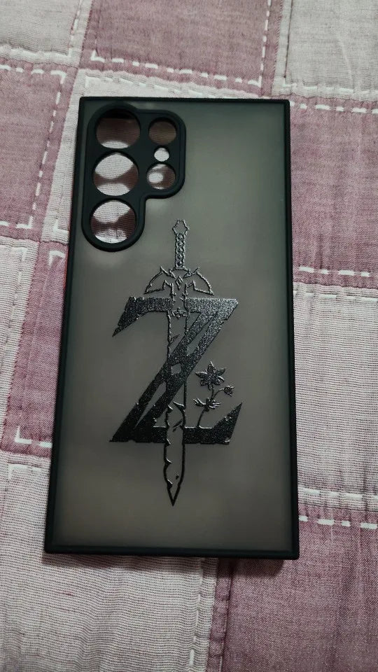 The Legend of Zelda phone case for Samsung, featuring a black sword and the letter "Z" design on a grey background reminiscent of the iconic Master Sword, lies on a patterned surface. The case includes precise cutouts for camera lenses and other phone features.