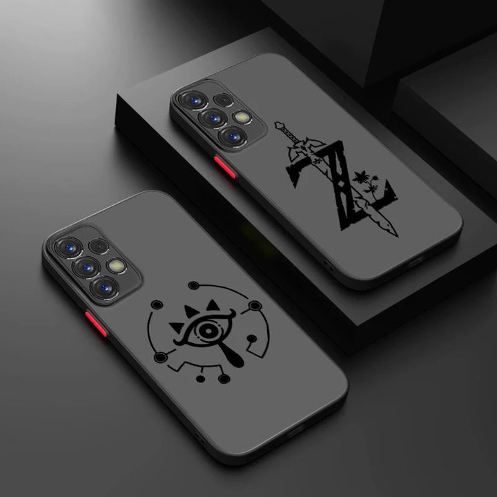 The image shows two grey smartphone cases from the Legend of Zelda for Samsung collection, each adorned with intricate black designs. One case features a stylized eye surrounded by elements that evoke the Triforce symbol, while the other displays a sword reminiscent of the Master Sword, intertwined with geometric patterns and stars. Both phones are equipped with multiple rear camera lenses.