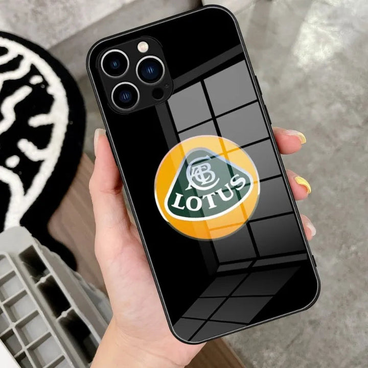 A person holds a smartphone equipped with the Lotus Glass for iPhone, which features a lightweight case embellished with a yellow and green Lotus emblem. The surface of the phone reflects the image of a window.