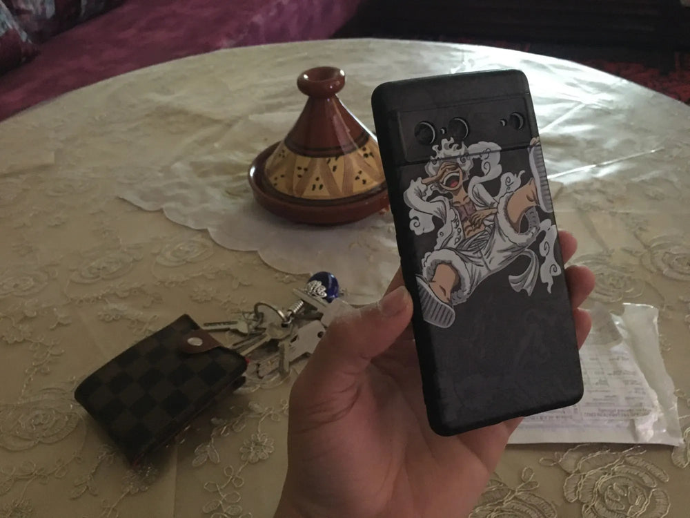 A hand holds a phone with the Luffy Gear 5 for Pixel case from the One Piece series, while keys and a wallet rest on a patterned tablecloth with a ceramic tajine in the background.