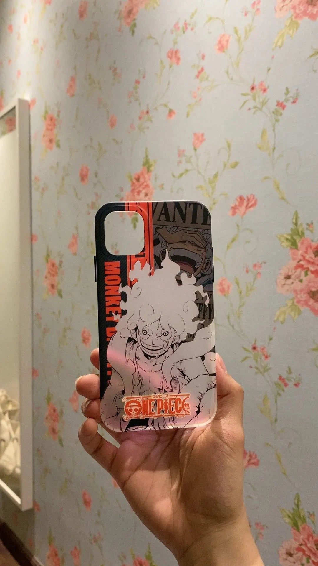 A hand holds a Luffy Gradient for iPhone case against a floral wallpaper background. This anti-scratch case ensures your phone stays protected while showcasing your favorite character in style.