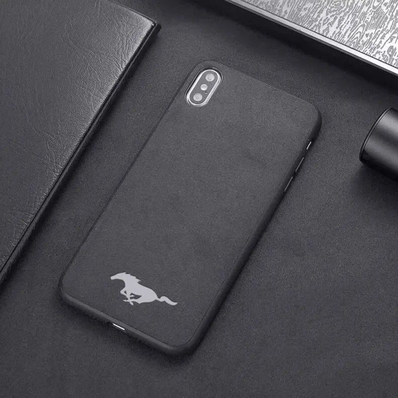 The Mustang for iPhone: A black smartphone case adorned with a white Mustang running horse logo on a black surface, pictured alongside a black notebook and other black items. This case boasts an anti-scratch coating and suede microfiber lining for enhanced protection.