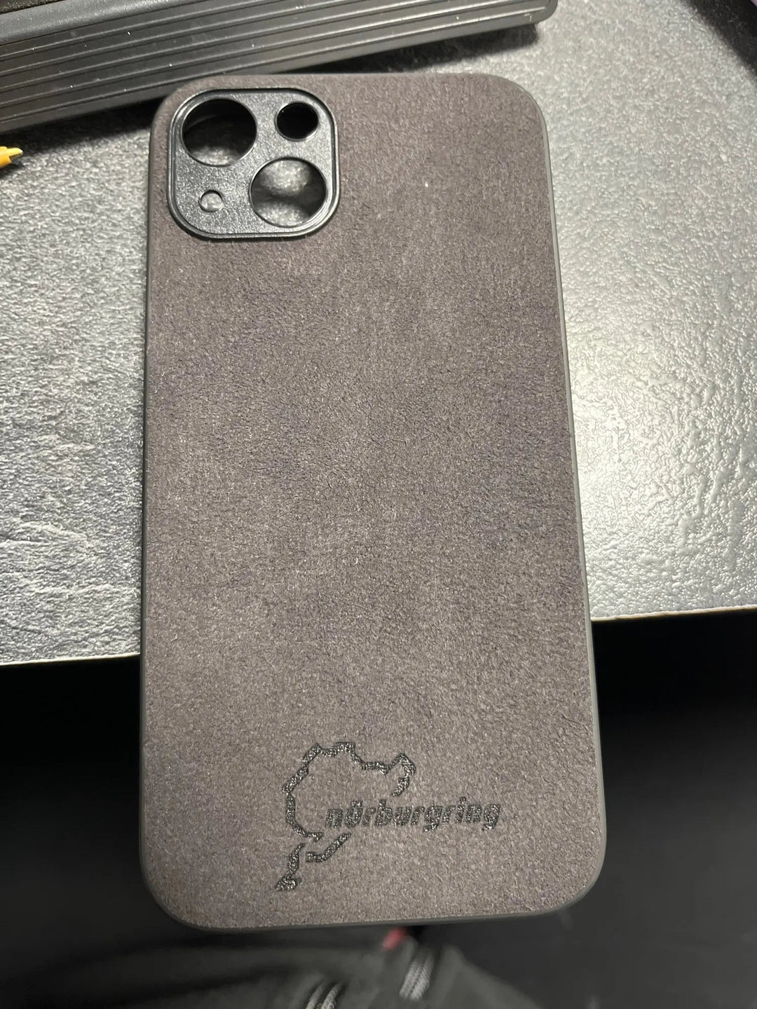 The Nurburgring for iPhone case, adorned with a Nurburgring logo and the iconic racing circuit design, rests on a dark surface. The sleek case is lined with suede microfiber for a soft touch and offers anti-scratch protection to keep your phone looking pristine.