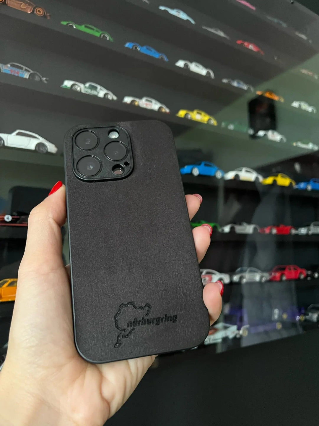 A hand holding the Nurburgring for iPhone, a black smartphone case featuring a small outline design and text, enhanced with anti-scratch protection. Multiple miniature car models are displayed on wall shelves in the background.