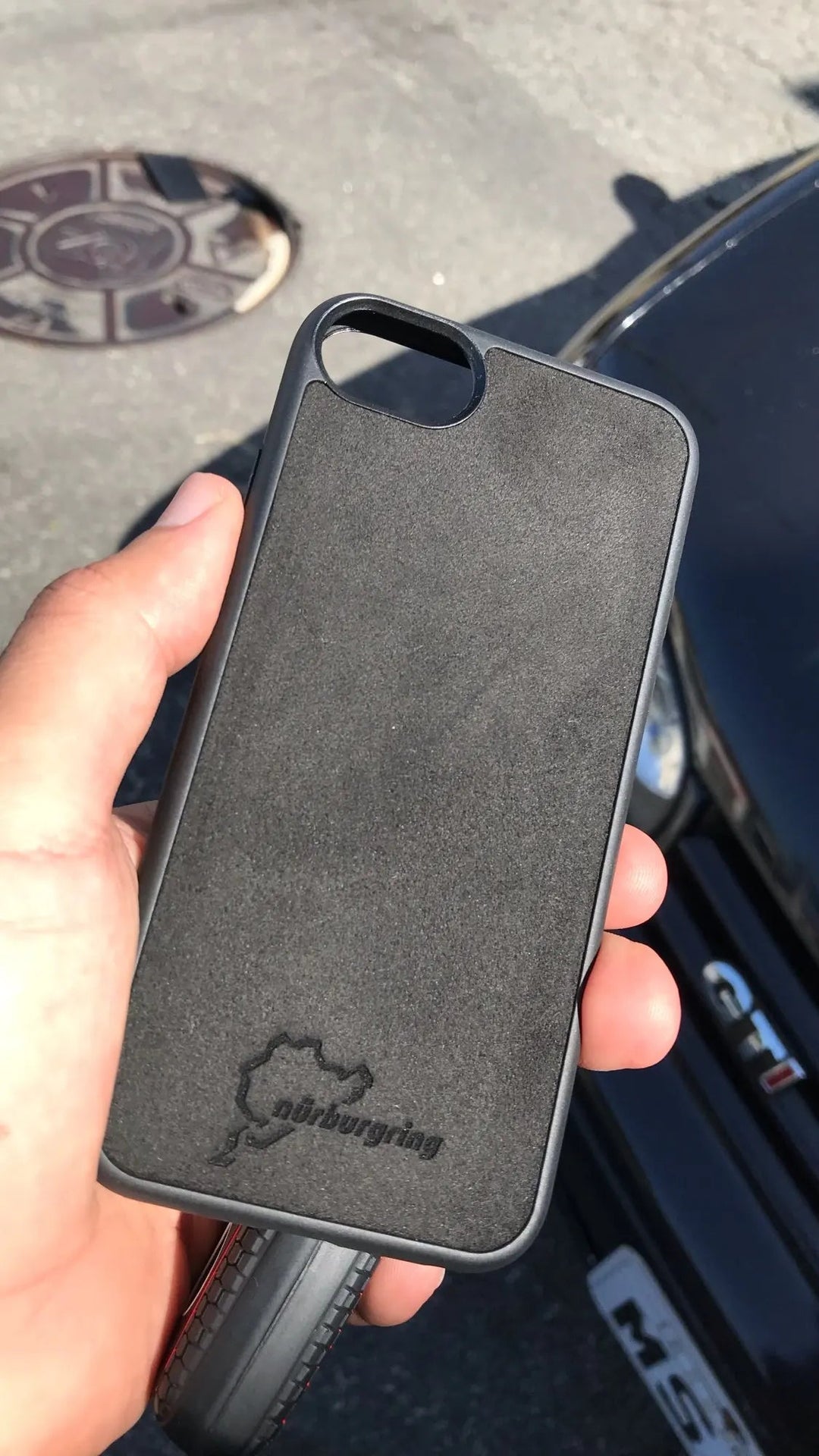 A person holds the Nurburgring for iPhone, a sleek black phone case emblazoned with the Nurburgring logo and crafted from anti-scratch suede microfiber. In the background, a car proudly displays its "GTI" badge.