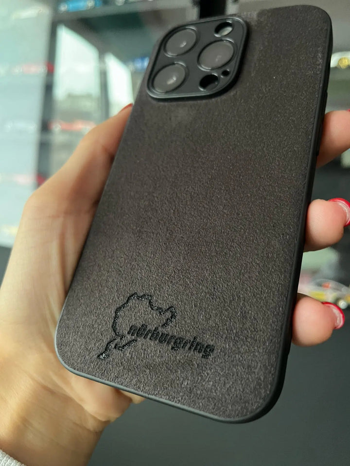 A hand holds an iPhone encased in the Nurburgring for iPhone, a dark, textured case adorned with the Nürburgring circuit outline and Nurburgring logo on the back, engineered from suede microfiber to ensure an anti-scratch finish.