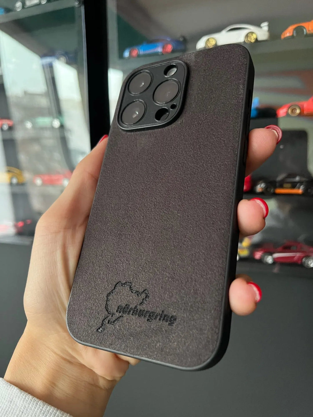 A hand holding an iPhone encased in the Nurburgring for iPhone, an anti-scratch, textured black cover adorned with the Nürburgring logo design. Model cars are displayed on shelves in the background.