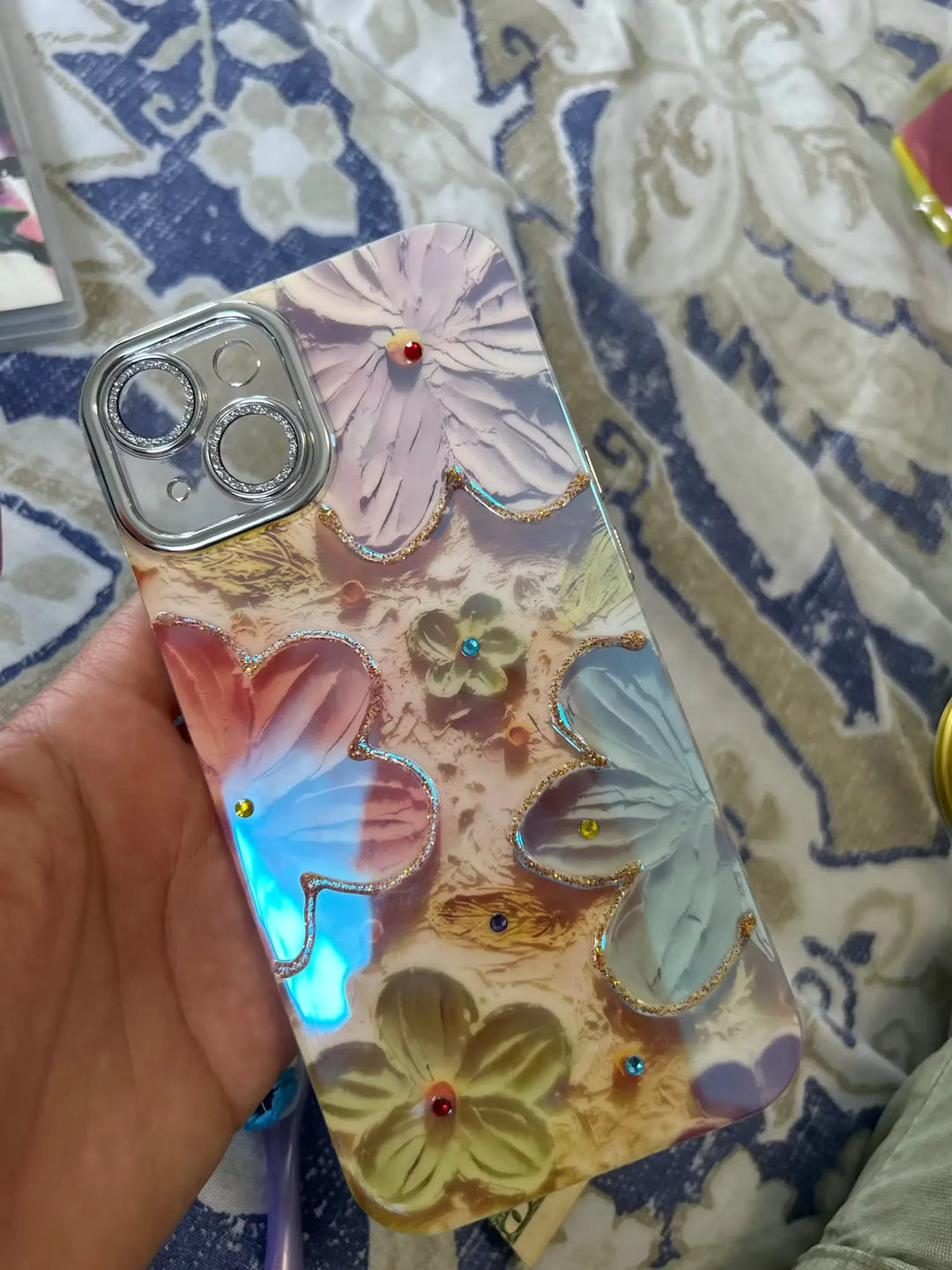 A close-up photo of a clear phone case with a 3D floral design. The flowers are painted in pastel colors and adorned with glitter and rhinestones. The case is being held in someone's hand.