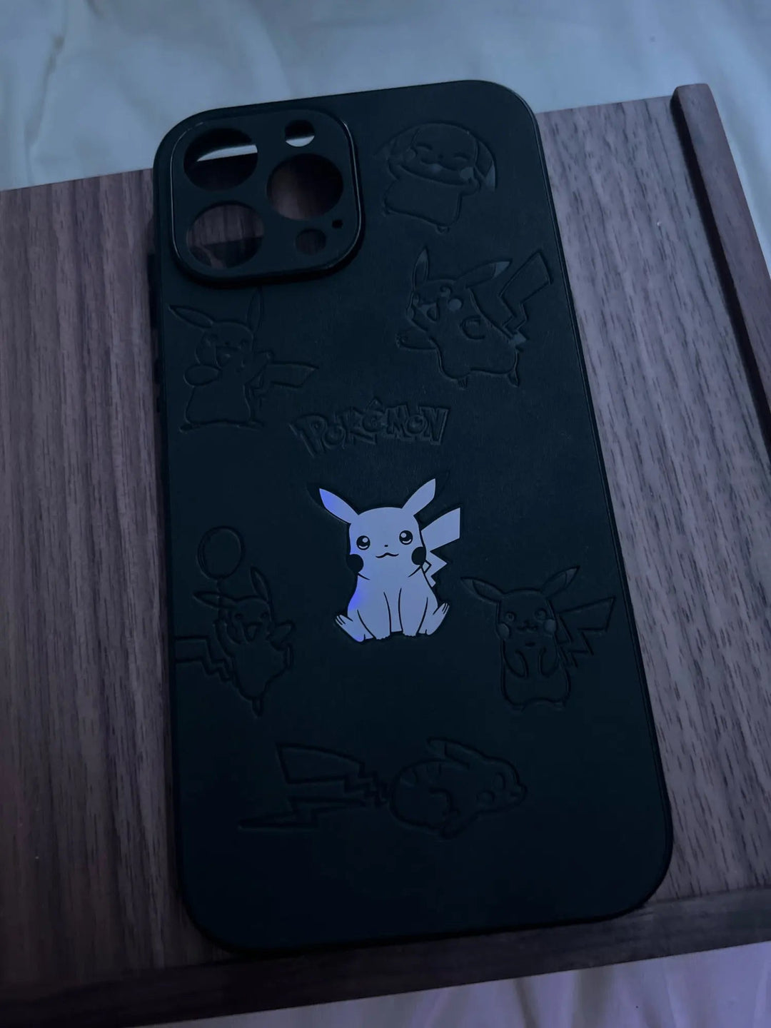 A black silicone phone case from the "Retro Pikachu for iPhone" collection is placed on a wooden surface, adorned with various etched Pokémon designs. The center features an illuminated Pikachu, bringing a retro-inspired design to life.