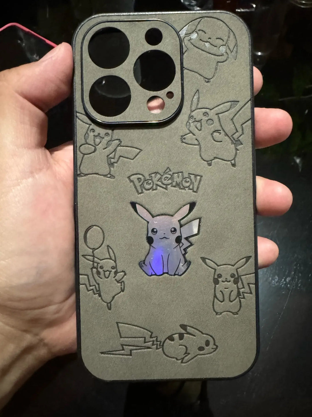 A hand holding the Retro Pikachu for iPhone, a silicone phone case adorned with various retro-inspired engravings of Pikachu in different poses. The case features one Pikachu illuminated in the center and has the word "Pokémon" engraved above the centered Pikachu.