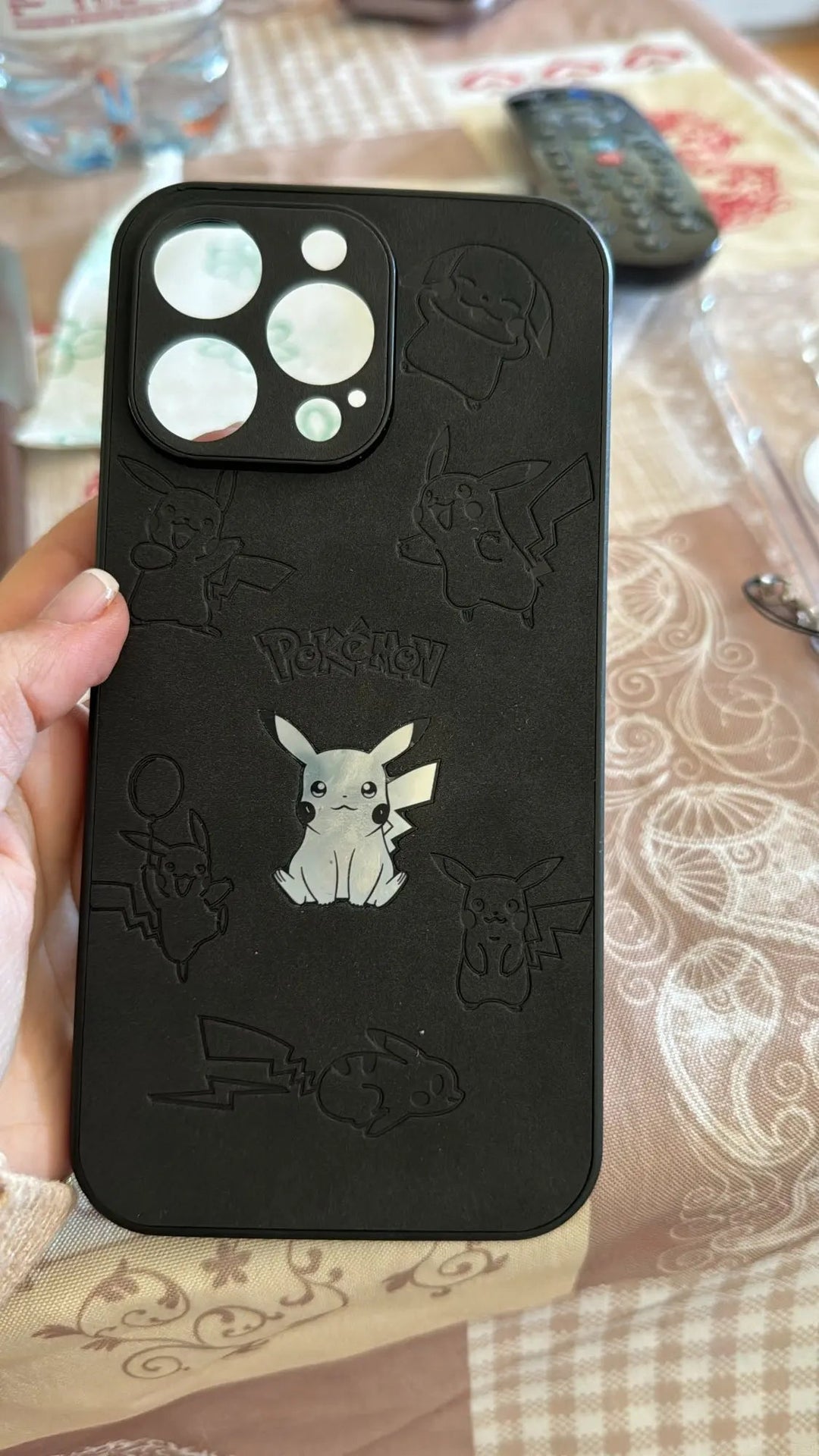 A close-up of the Retro Pikachu for iPhone showcases its retro-inspired black silicone design, featuring multiple engraved illustrations of Pikachu and the Pokémon logo. The case is being held in hand against a background that includes a patterned tablecloth and various items.