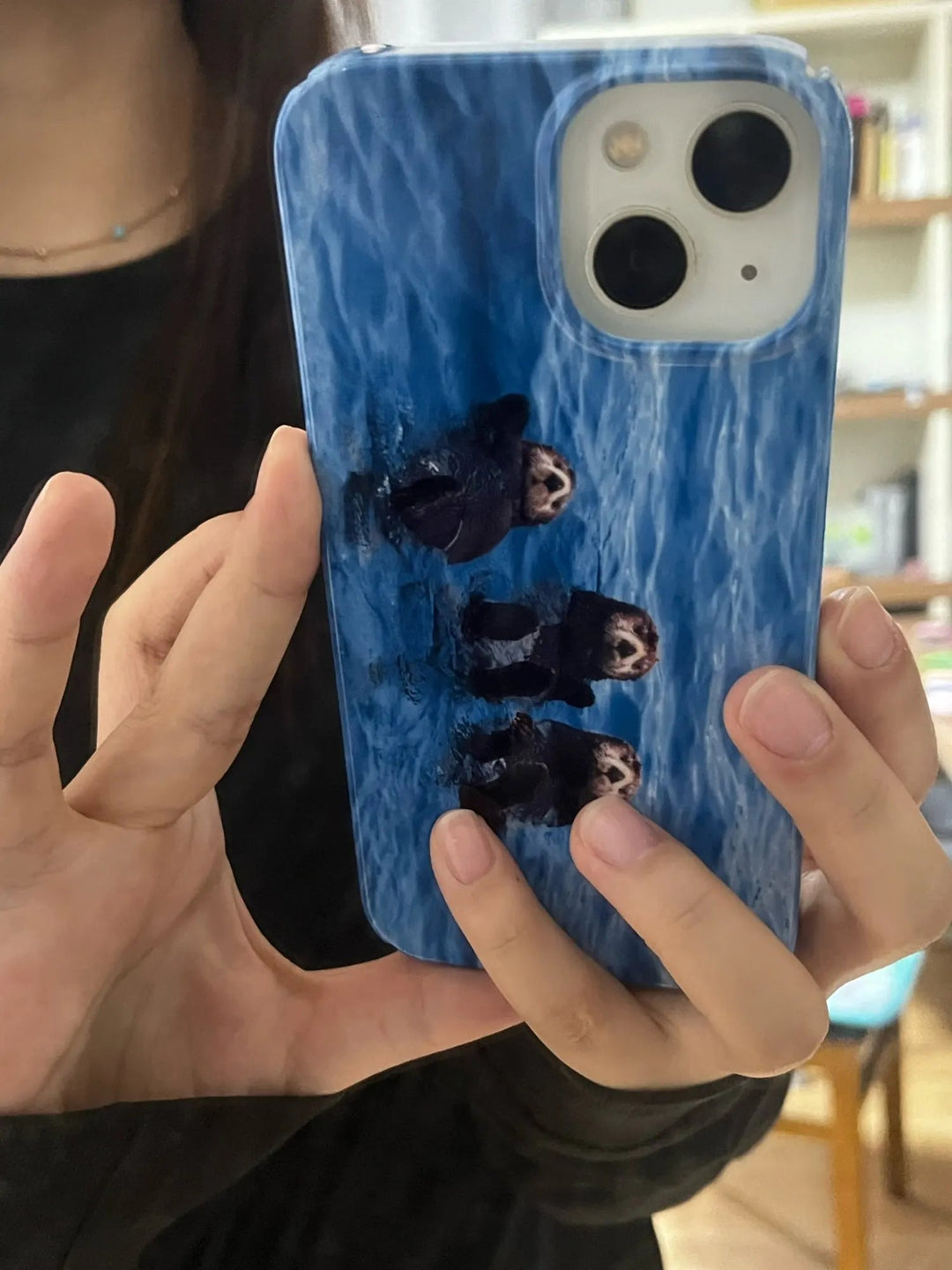 A person holding an iPhone protected by the Sea Otter Case - Acrylic, which features a blue backdrop with an enchanting image of three otters swimming, ideal for animal enthusiasts.