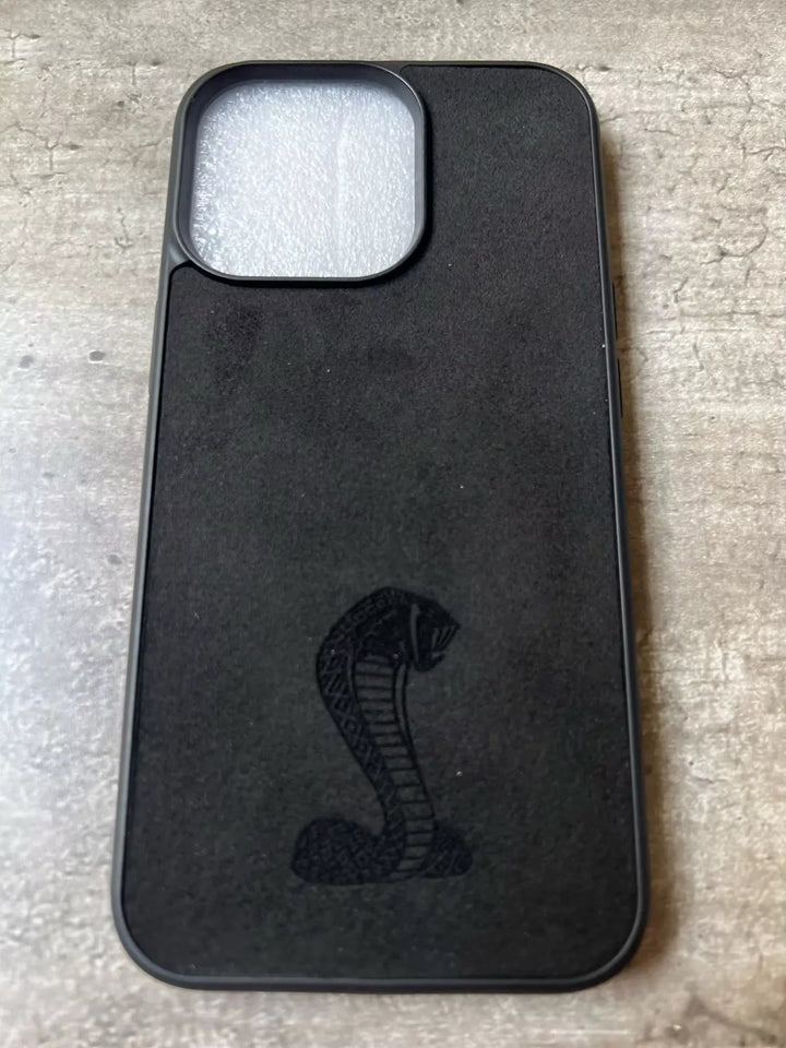 The Shelby for iPhone, a black phone case, showcases an embossed cobra emblem on a textured surface against a stone-like background. This Anti-Scratch accessory adds a touch of automotive heritage to your gear.