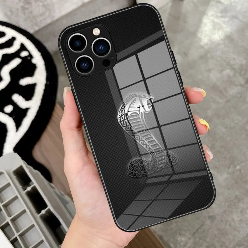 A hand holds the Shelby Glass for iPhone, featuring a tempered glass case adorned with the Shelby Cobra emblem, reflecting a segmented window pattern. In the background, a black and white patterned mat is visible.