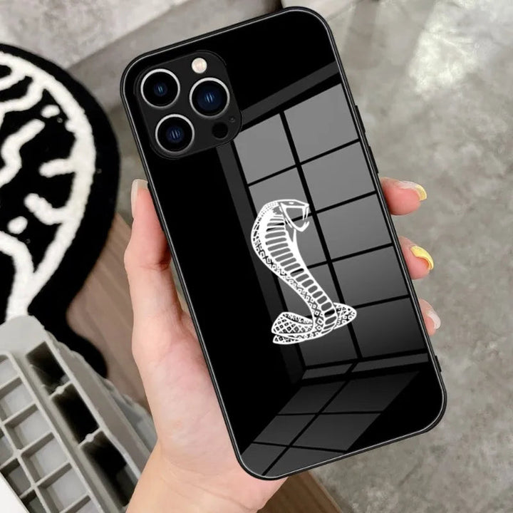 A hand holding a black iPhone encased in the Shelby Glass for iPhone, which features a white Shelby Cobra emblem on the back. The background reveals a reflective floor and a geometric patterned rug.