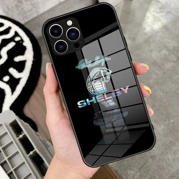 A hand holding a black smartphone with the Shelby Glass for iPhone, featuring the Shelby Cobra emblem and a sleek cobra design.