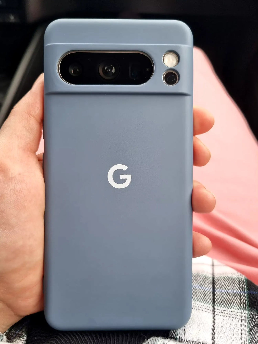 Close-up of a hand holding a Google Pixel smartphone with a "G" logo on the back, encased in a sleek Silicone Case for Google Pixel. The phone features a prominent camera module with multiple lenses and a flash. The person is dressed in a plaid shirt and pink pants.