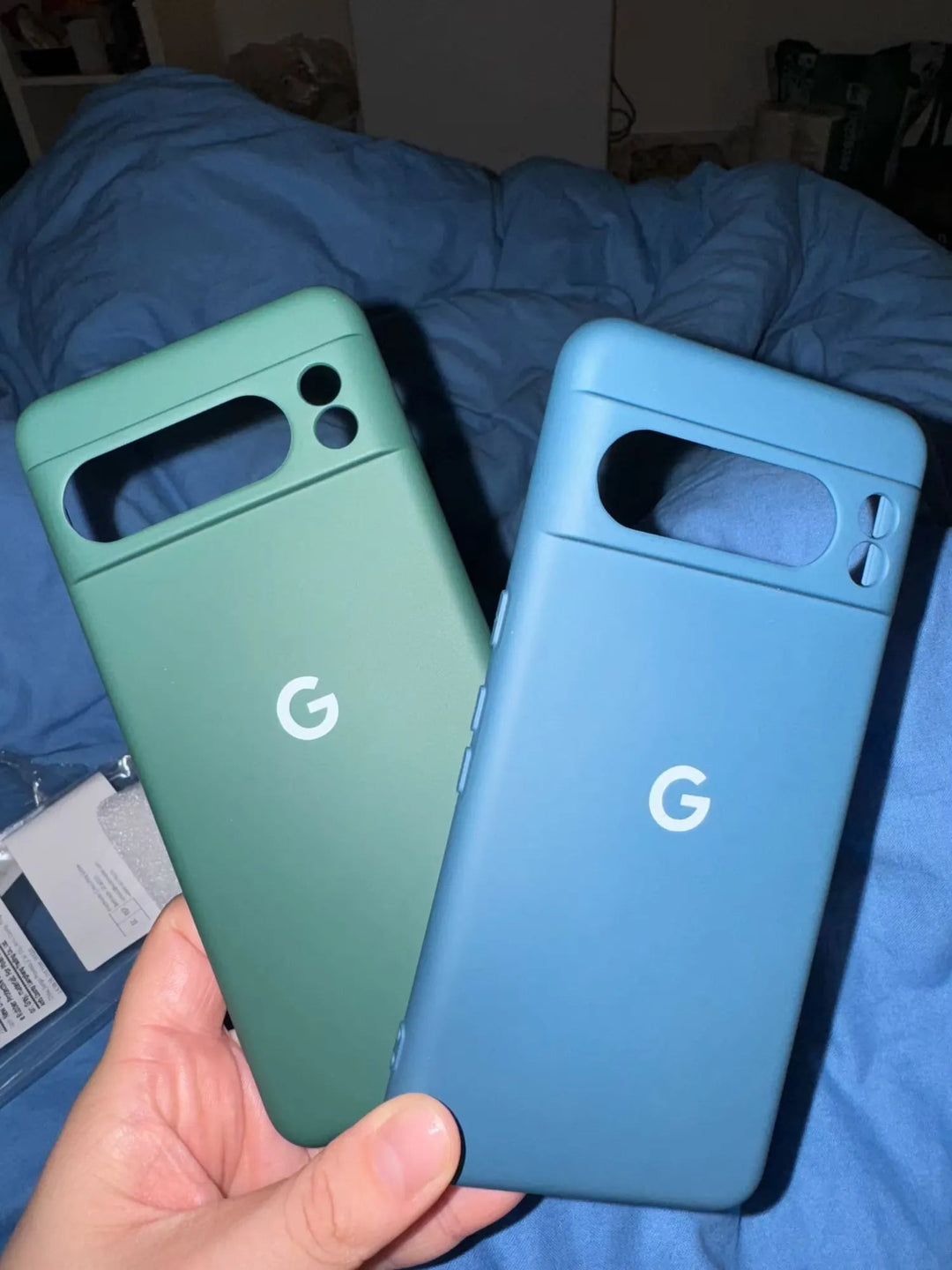 A hand holds two slim-fit Silicone Cases for Google Pixel, one in green and the other in blue, against a background of a blue blanket.