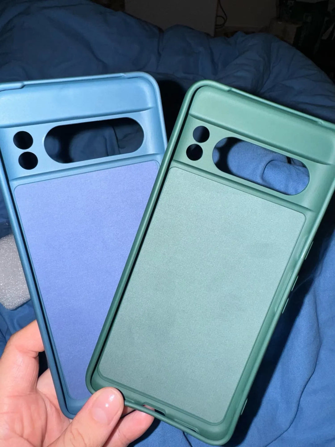 Holding two slim-fit Silicone Cases for Google Pixel, one in blue and the other in green, each featuring a prominent cutout at the top flanked by two smaller ones. Set against a blue fabric backdrop, these cases are designed to provide maximum protection and anti-scratch features to keep your device pristine.