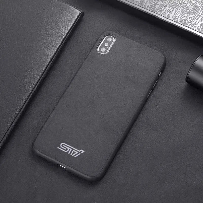 A sleek and stylish black smartphone with a minimal rear camera design sits on a black surface next to a black notebook, a black pen, and a black cylindrical object. The STI for iPhone case adds an extra touch of elegance to the scene.