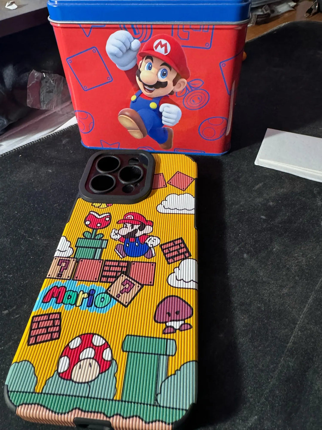 The Super Mario for iPhone is placed on a dark surface in front of a blue and red Super Mario-themed tin box. The hard plastic case offers both stylish design and protective functionality.