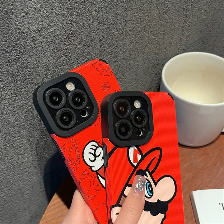 Two "Super Mario for iPhone" smartphones encased in red Super Mario design covers are held in a hand. A white cup and a book are on the table in the background.