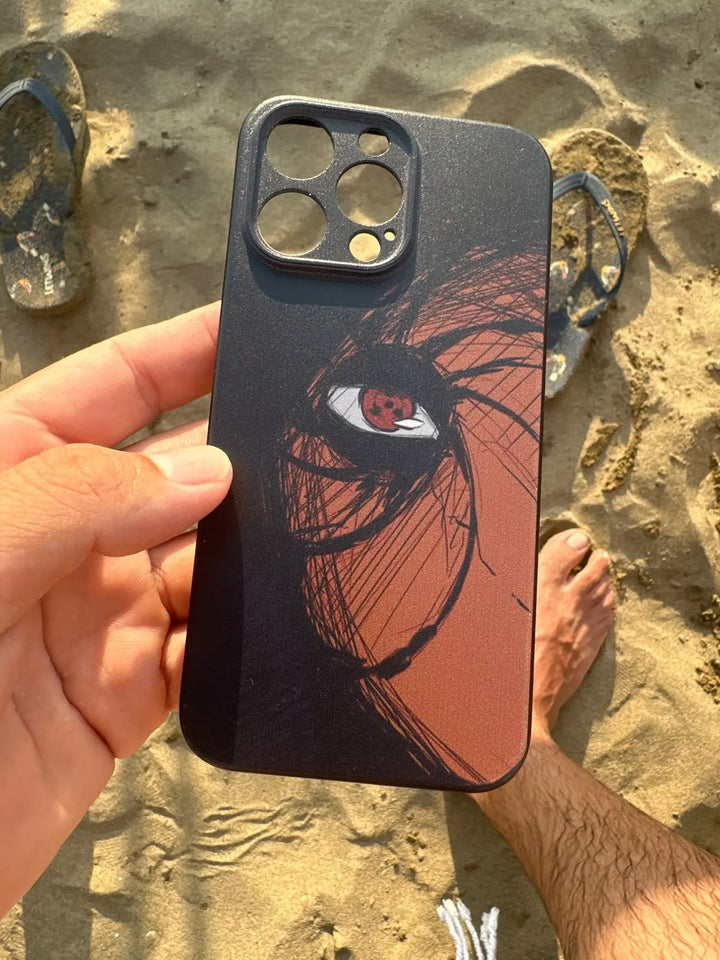 A hand holds the Tobi Mask for iPhone, a sleek black TPU case adorned with an artistic depiction of a character's eye and a swirling mask design, while sandy flip-flops and a bare foot rest on the beach in the background.