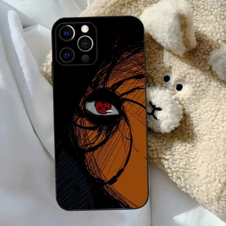 A Tobi Mask for iPhone, which is a black TPU case showcasing an artistic design with a close-up of a red-eyed character and a swirling mask pattern, sits beside a beige, plush bear toy on a white bedspread.